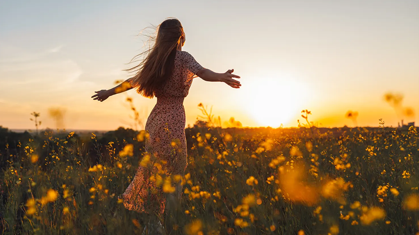 Person with outstretched arms, walking through a field of flowers at sunset